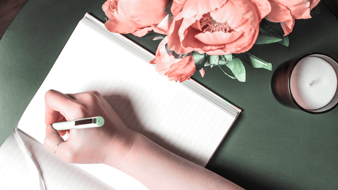 A dark green desktop featuring a woman's arm writing in an open journal. Adjacent to the journal, there are pink flowers and a jar candle, creating a serene and inviting workspace.