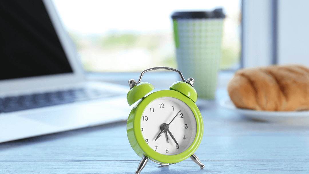 An illustrative image for a blog post about daily routines. In the foreground, a lime green alarm clock takes center stage on a well-kept desk, symbolizing the importance of time management in one's daily routine. In the background, a laptop, a delicious croissant, and a light blue disposable coffee cup hint at the elements that make up a morning ritual.