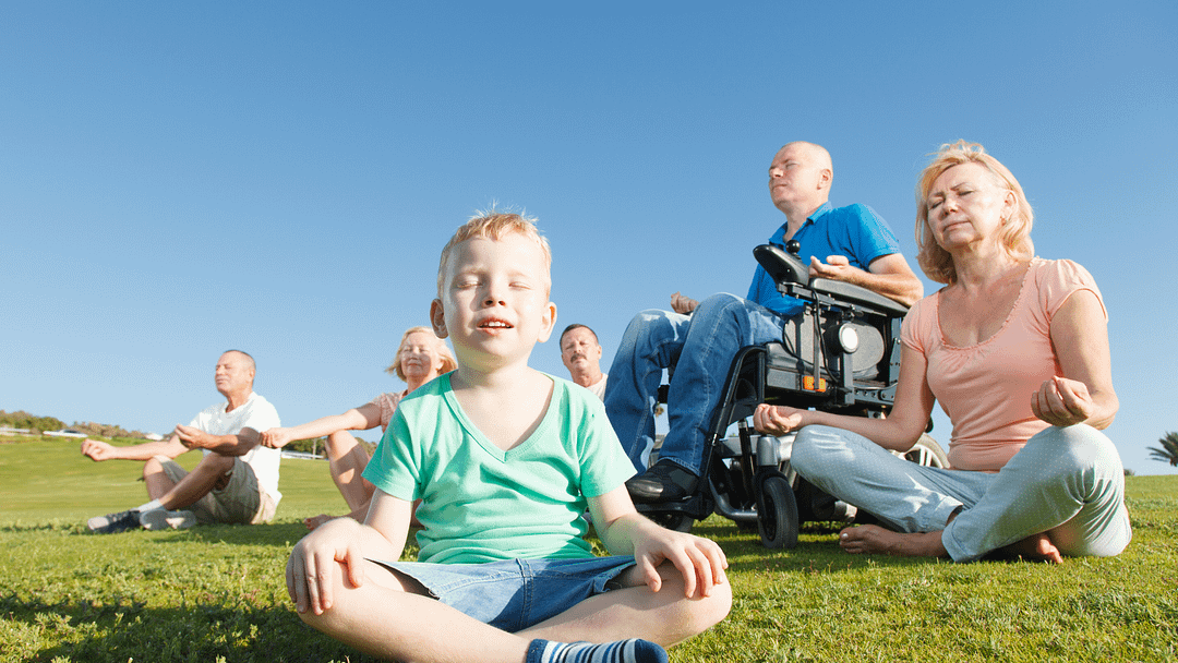 Discover 5 low-energy family time ideas for chronic illness warriors. | Image ID: A multi-generational family meditating on the grass on a sunny day. One of the adult males is in a motorized wheelchair.